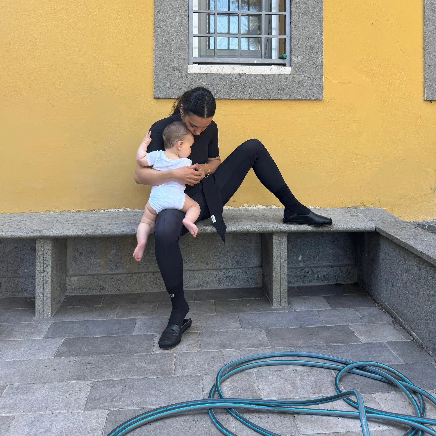 Mamma con leggings neri e scarpe comode abbraccia neonato su panchina gialla